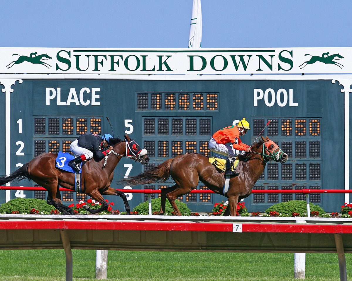 Two horses racing past the green Suffolk Downs tote board